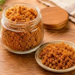 Toples kaca berisi abon sapi berwarna cokelat keemasan dengan seporsi abon di piring kecil di atas meja kayu.