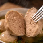Close-up bakso sapi yang dibelah menampilkan tekstur daging yang padat dan kenyal dengan latar kuah bakso panas yang mengepul.