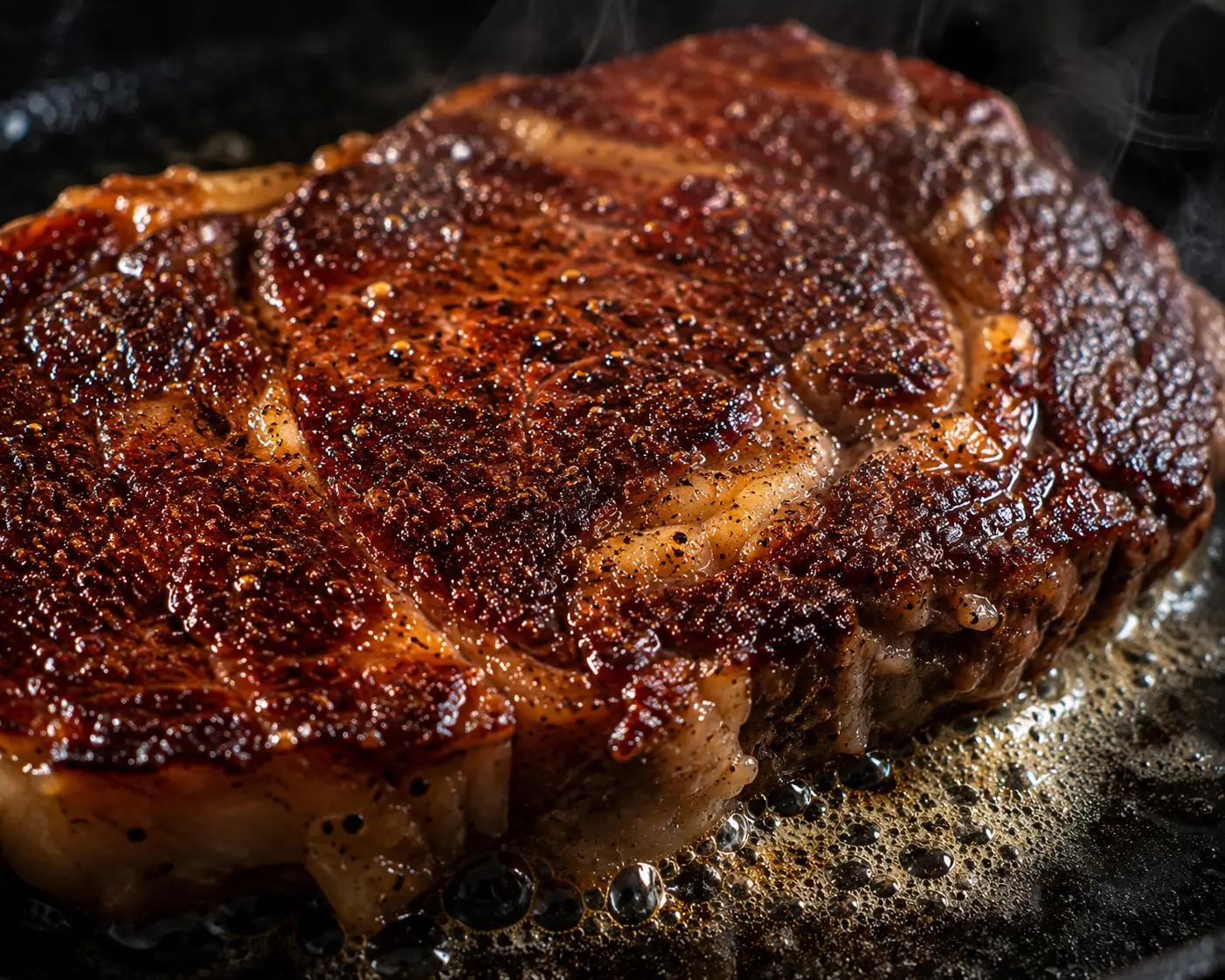 Close-up kerak cokelat gelap pada steak ribeye yang sedang di-sear di atas cast iron dengan minyak berbuih dan asap tipis.