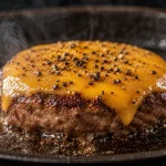 Beef patty tebal dipanggang di wajan cast iron dengan lelehan keju cheddar, lada hitam kasar, dan kepulan asap yang memperlihatkan tekstur juicy.