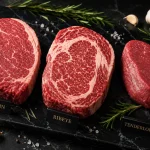Potongan daging steak Sirloin, Ribeye, dan Tenderloin mentah di atas nampan marmer hitam dengan rosemary, bawang putih, dan garam laut.
