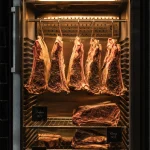 Bagian dalam dry aging chamber dengan potongan daging sapi yang digantung dan kontrol suhu digital.