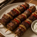 Tiga skewer beef kebab panggang dengan daging sapi kecokelatan, paprika merah, bawang bombay panggang, dan saus garlic yogurt di atas piring putih.