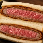 Potongan wagyu katsu sando dengan daging merah muda, lapisan tepung renyah, dan roti putih lembut.